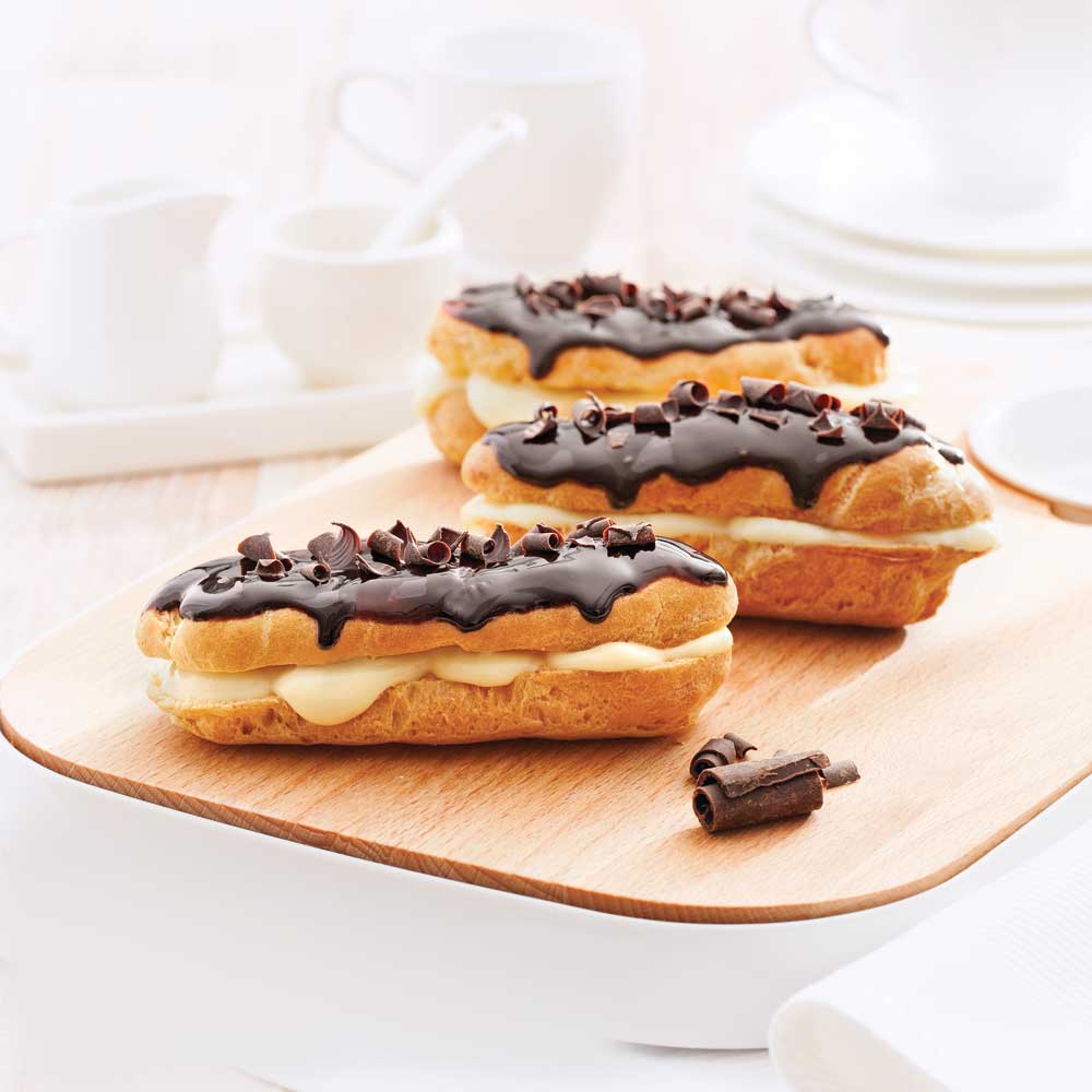 Trois éclairs au chocolat: pâte à choux garnie de crème pâtissière et ganache au chocolat sur le dessus sur une planche de bois.
